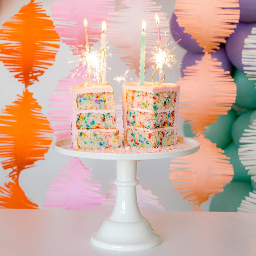 Sparkler candles on top of a funfetti cake with colorful garlands in the background, and other party supplies.