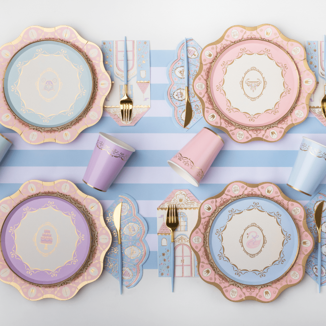 A styled flat-lay of pastel party tableware on a white surface with light blue and white stripes. The set includes scalloped plates in pink, lavender, and light blue with gold detailing, matching cups, napkins, and gold-toned cutlery.