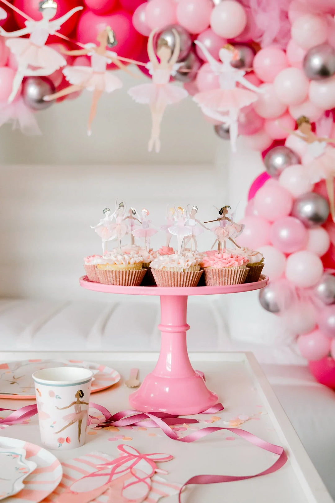 Ballerina birthday dessert table with ballerina-topped cupcakes on a pink cake stand beneath a pink balloon garland with hanging ballerina figures