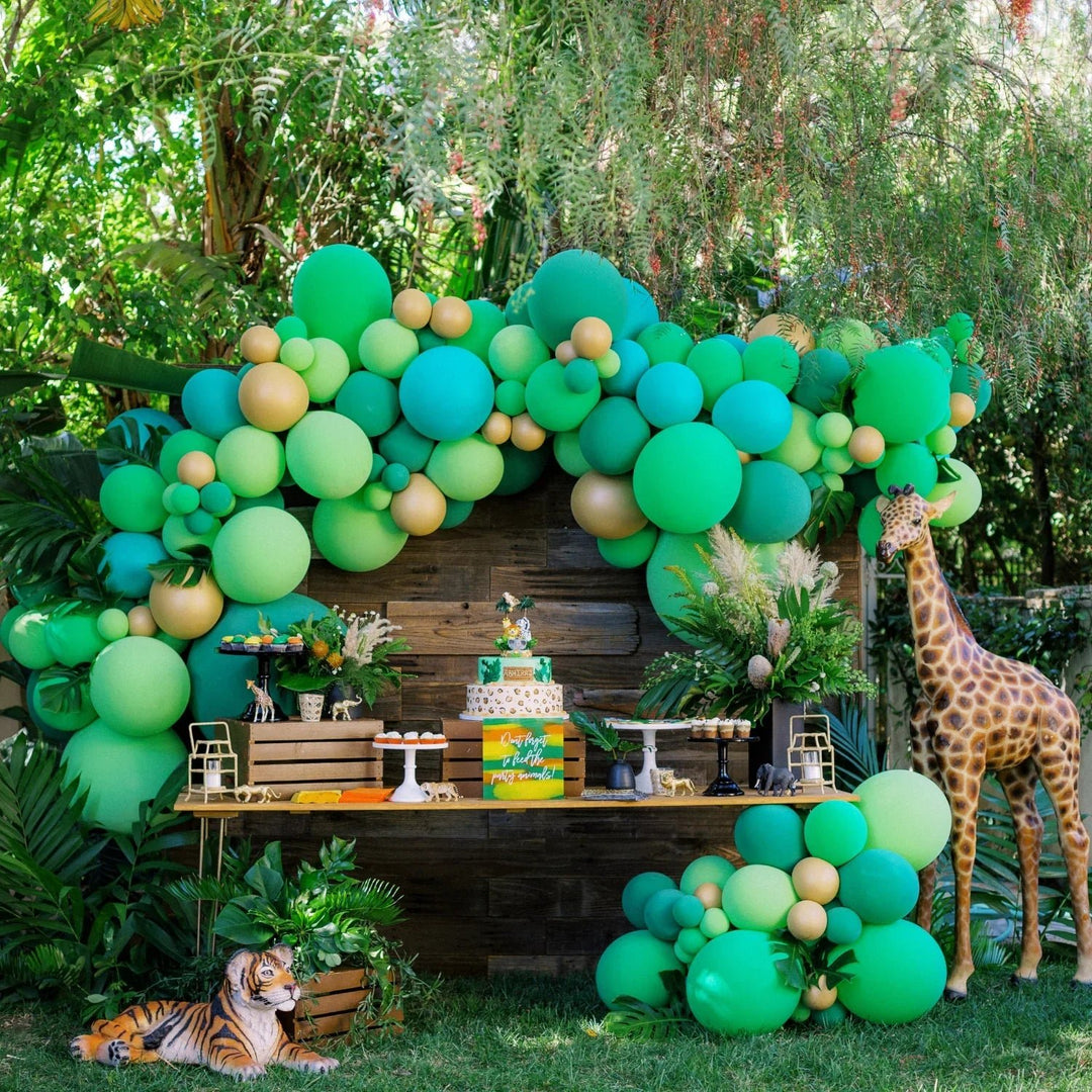Outdoor safari dessert table with a green and gold balloon garland, safari cake, giraffe and tiger statues, and lush greenery