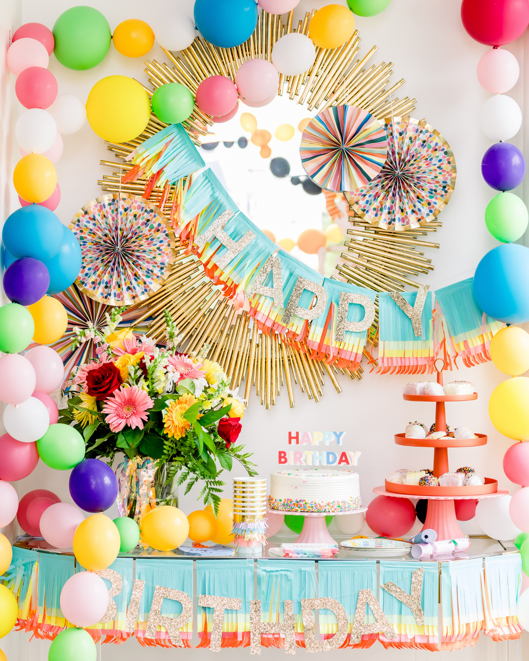 Birthday dessert table with a rainbow balloon garland, a fringe Happy Birthday banner, paper fans, and a sprinkle cake