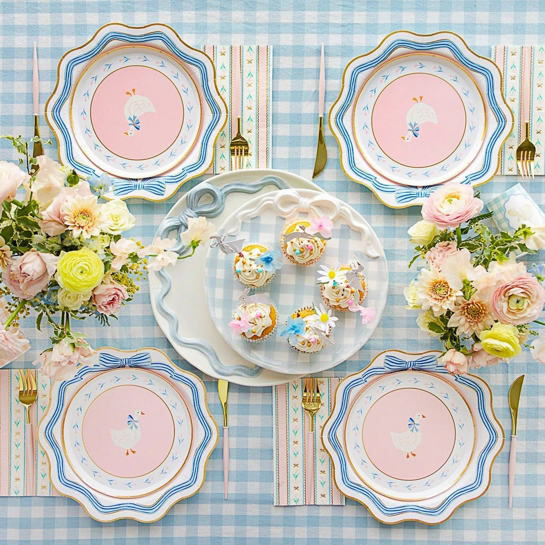 Blue gingham Easter table with goose print plates, a bow platter of cupcakes, and pastel flower bouquets