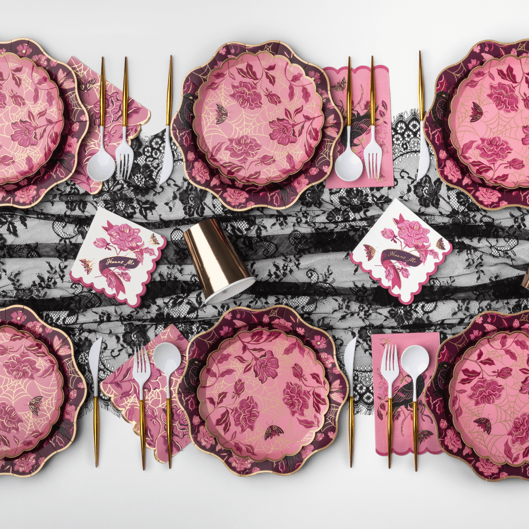A styled flat-lay of a pink and magenta floral tableware set with gold-toned cutlery, white napkins, and a black-and-white lace runner on a light gray background.