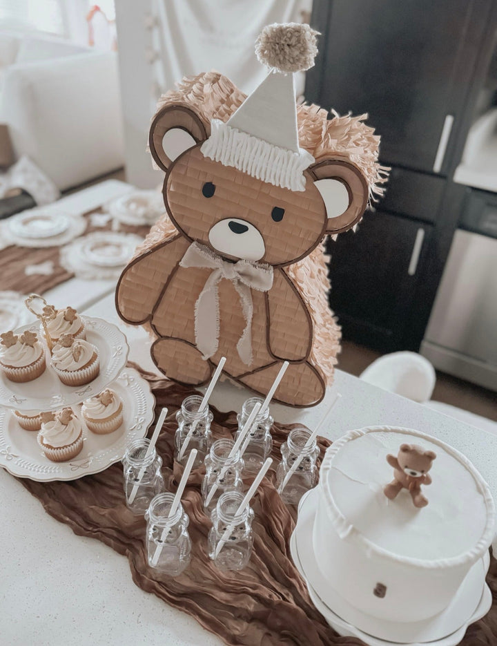Teddy bear piñata styled behind a white cake, cupcakes, and milk bottles on a teddy bear party table