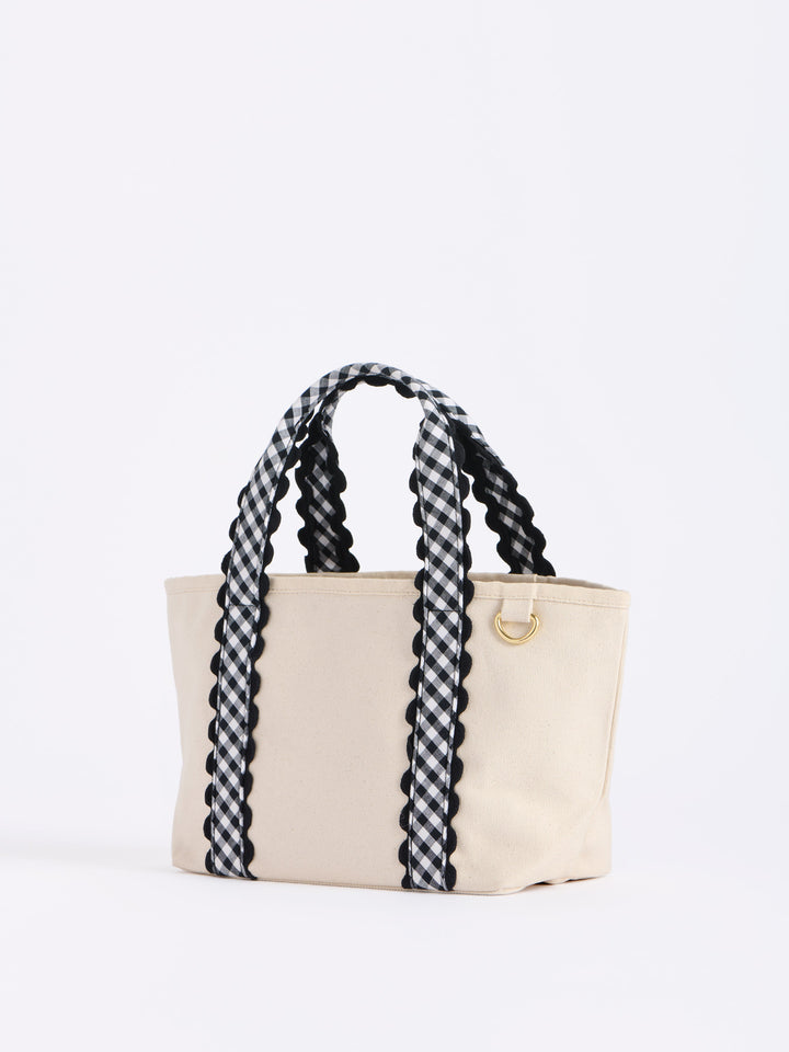 A cream canvas tote bag with thick black and white checkered braided shoulder straps and a small gold metal ring detail on the side, photographed against a plain white background.