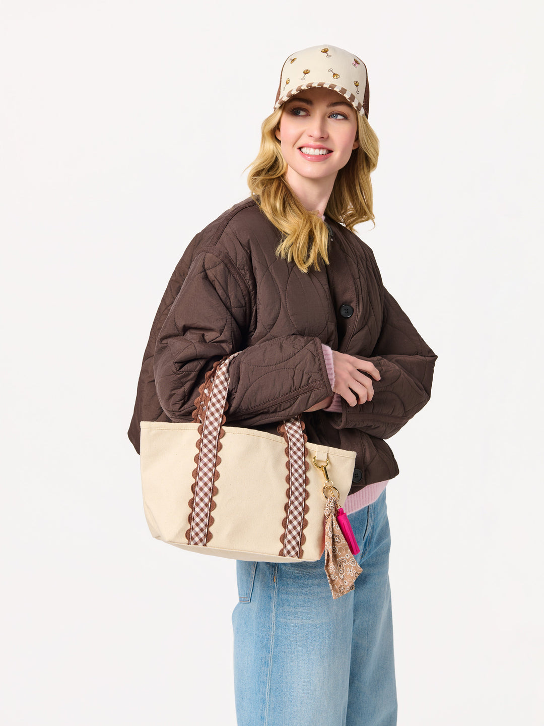 A woman modeling a brown quilted puffer jacket, light blue jeans, a beige baseball cap with brown stars, and carrying a beige tote bag with brown patterned straps, against a plain white studio background.
