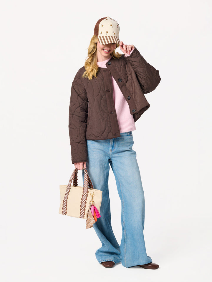 A woman models a brown quilted cropped jacket, styled with a pink t-shirt, wide-leg blue jeans, a beige and brown patterned cap, and a matching beige tote bag with pink tassels, set against a plain white studio background.