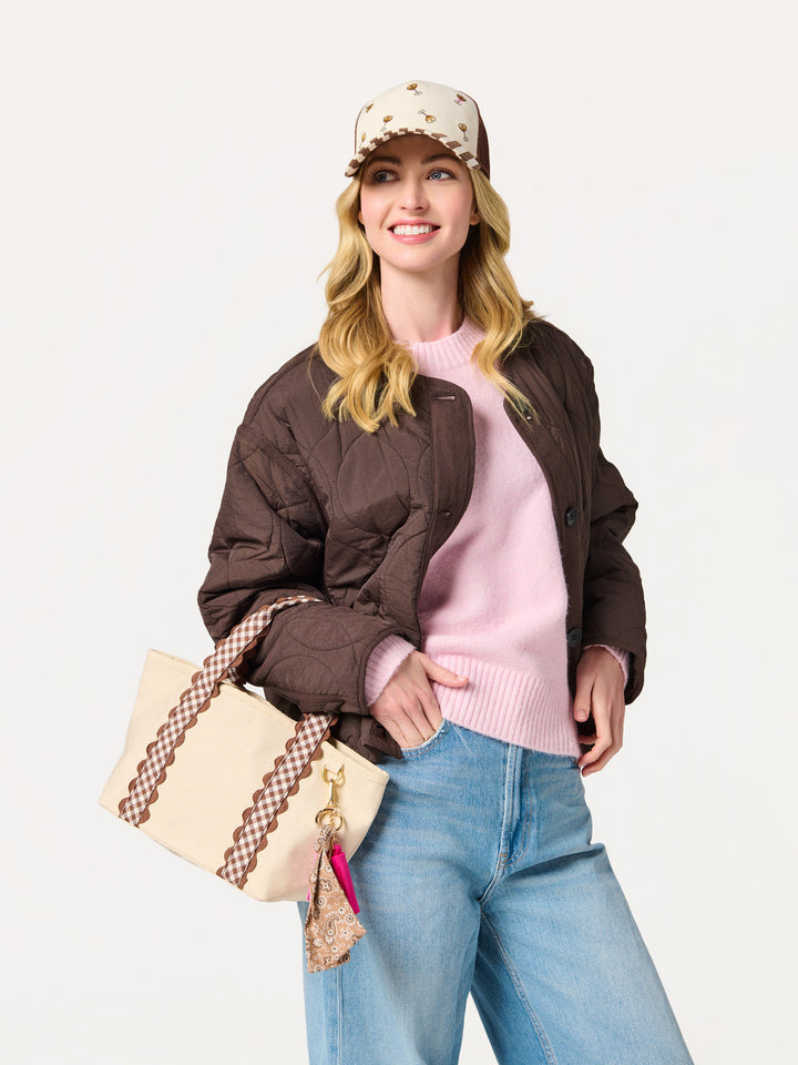 A woman modeling a dark brown quilted bomber jacket over a light pink sweater, paired with blue jeans, a beige patterned baseball cap, and a cream tote bag with brown checkered straps, against a plain white studio background.