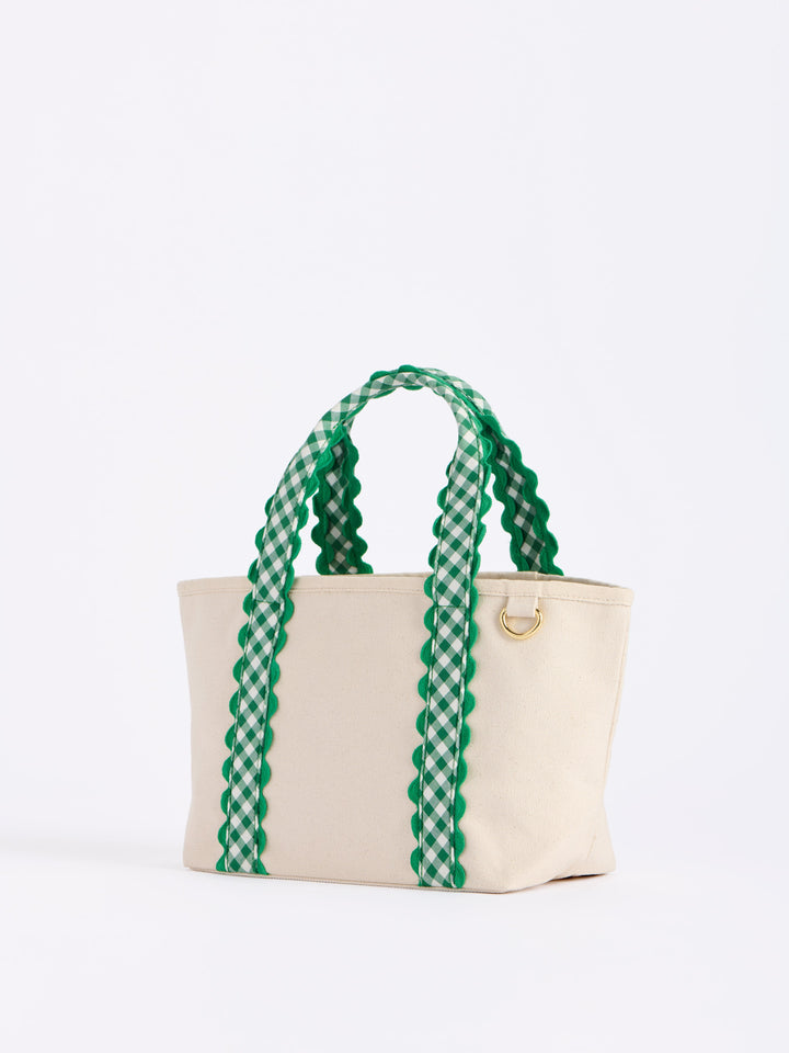 A cream canvas tote bag with green and white gingham patterned handles and a small gold metal ring detail, photographed on a clean white background.