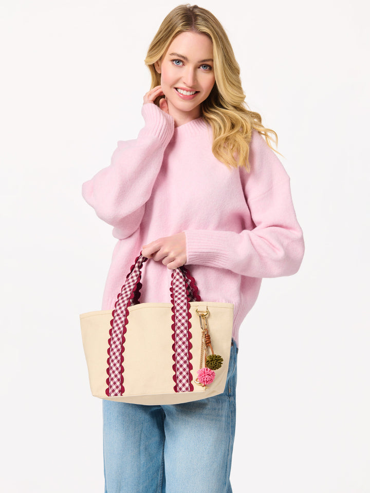 A woman wearing a pink sweater and blue jeans holds a cream canvas tote bag with red-and-white patterned handles and a pink flower charm, against a plain white background.