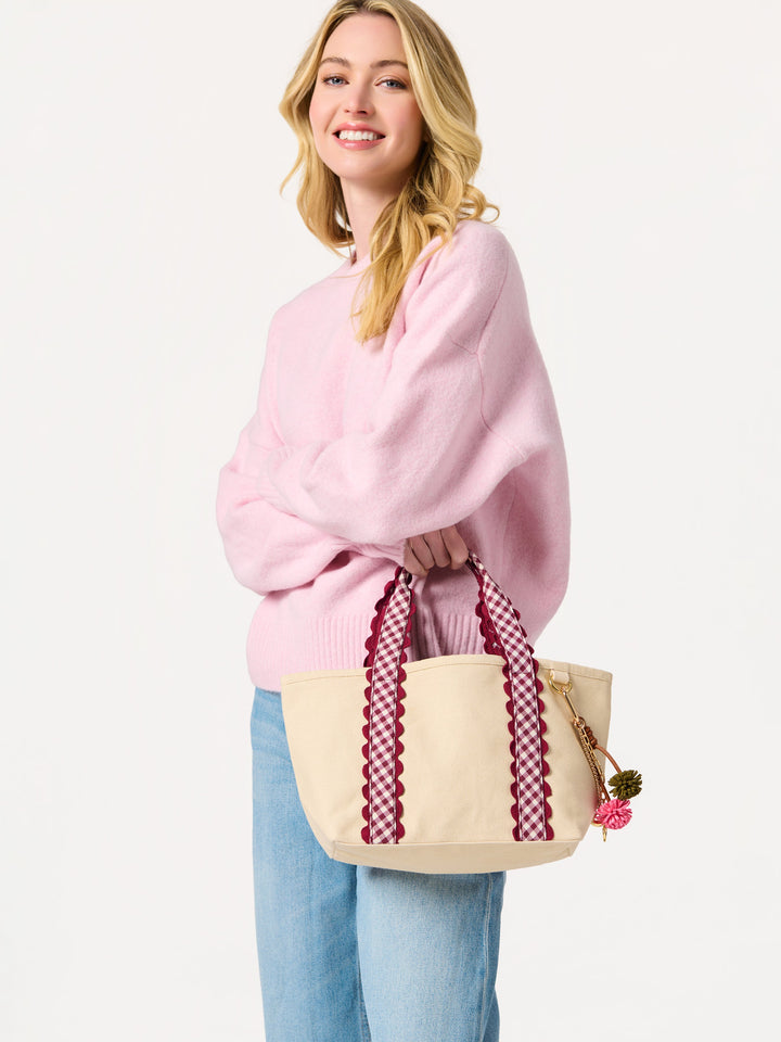 A woman wearing a pink sweater and blue jeans holds a cream canvas tote bag with burgundy patterned handles against a plain white background.