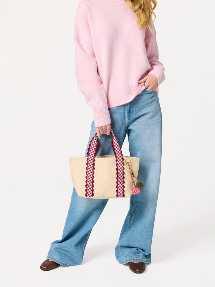 A model wearing a pink sweater, blue jeans, and brown shoes holds a beige tote bag with red-and-white patterned handles against a plain white studio background.
