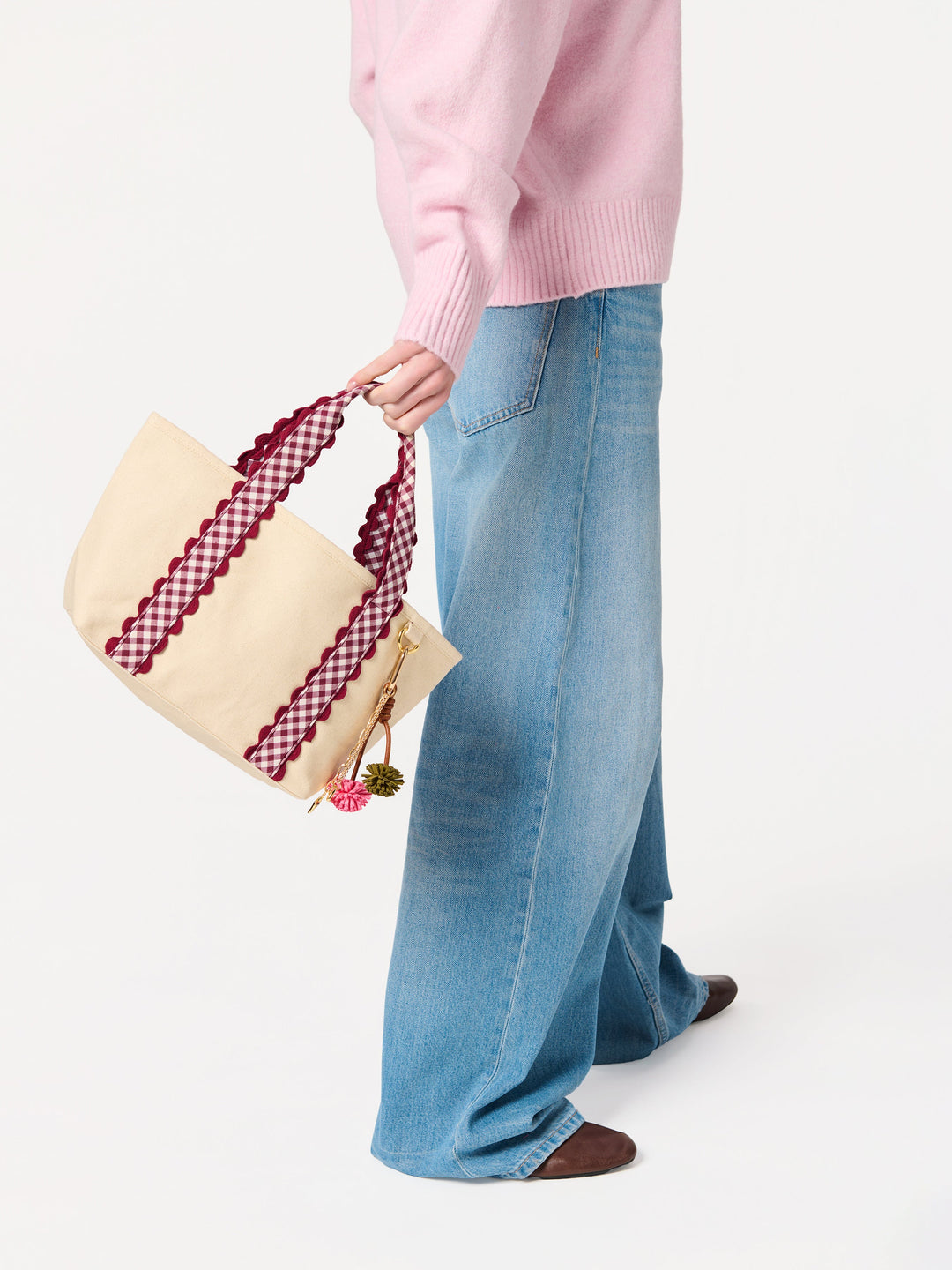 A person wearing a pink sweater and wide-leg blue jeans holds a cream canvas tote bag with burgundy and white gingham patterned shoulder straps against a plain white background.