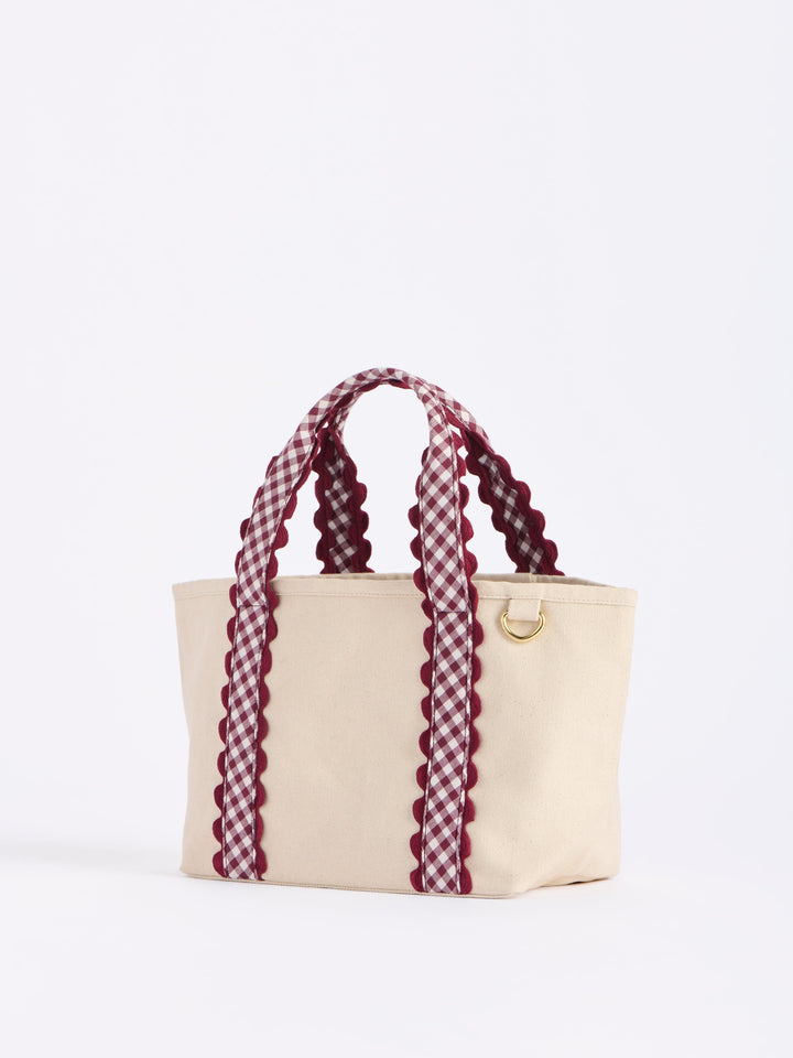 A cream canvas tote bag with burgundy and white gingham patterned handles and side trim, featuring a small gold metal ring on the front.