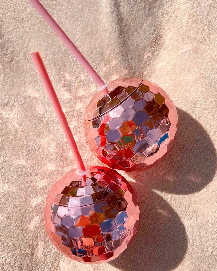 Two pink disco ball cups with straws casting shadows on a sandy blanket