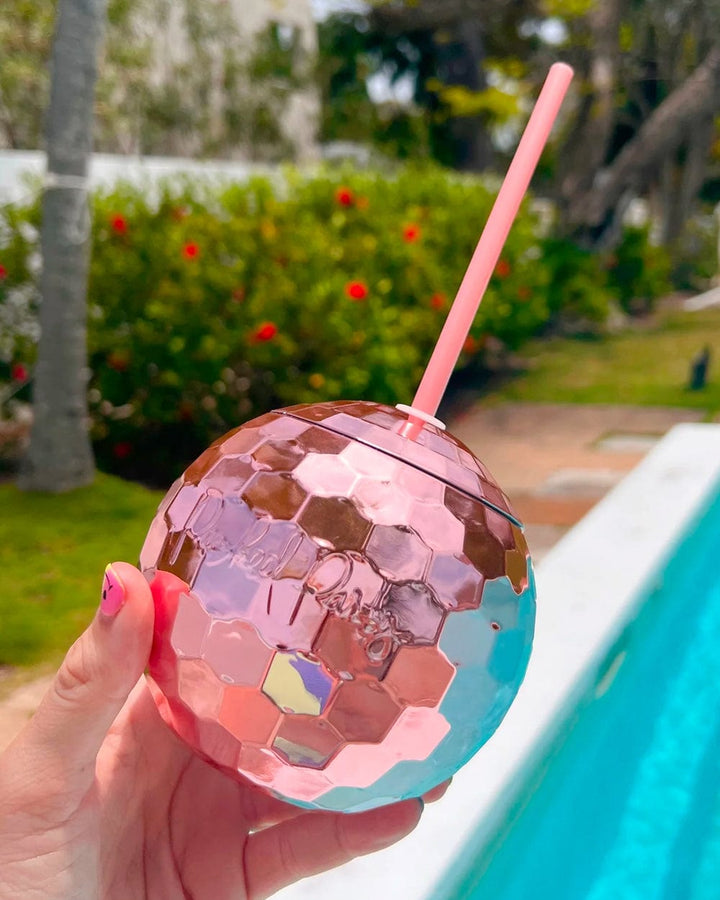 Hand holding a pink disco ball cup beside a swimming pool and flowering hedge
