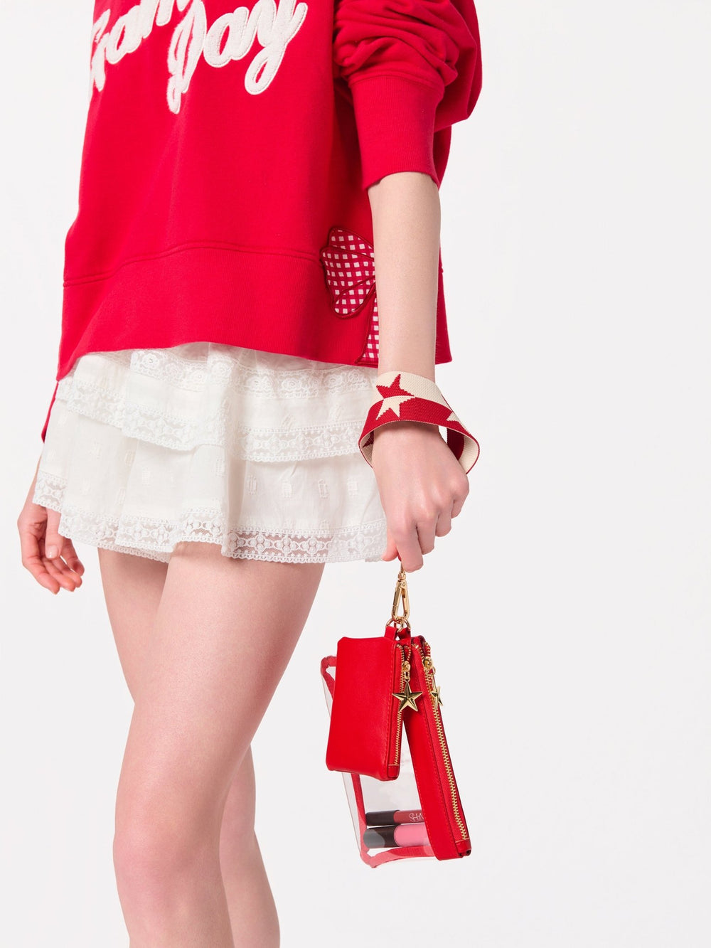 A model wearing a red sweatshirt and white lace skirt holds a red phone pouch with a clear side panel and gold star charm against a plain white background.