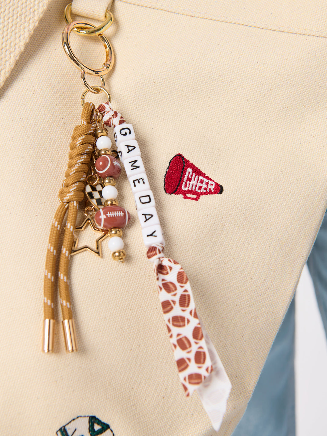A close-up of a beige canvas tote bag featuring a custom football-themed keychain charm. The keychain includes a gold-tone split ring, a braided tan rope tassel, football-shaped beads, a white letter tile spelling 'GAME DAY', and a white ribbon printed with brown football patterns and a red 'CHEER' megaphone patch.