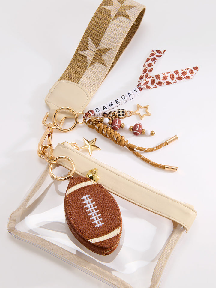 A clear plastic pouch with a beige zipper and strap, decorated with a brown football-shaped charm, gold keychain rings, and a patterned wristlet strap on a white background.