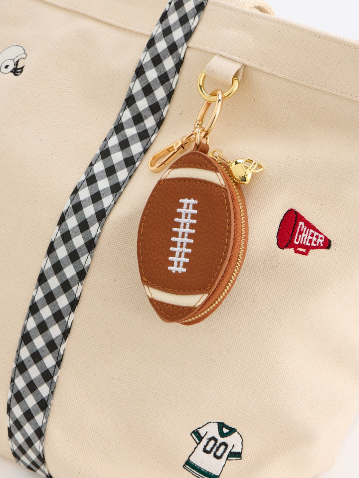 Close-up of a beige canvas tote bag with a black-and-white gingham strap, a brown football-shaped zipper pouch with gold hardware, and small embroidered cheer-themed patches.