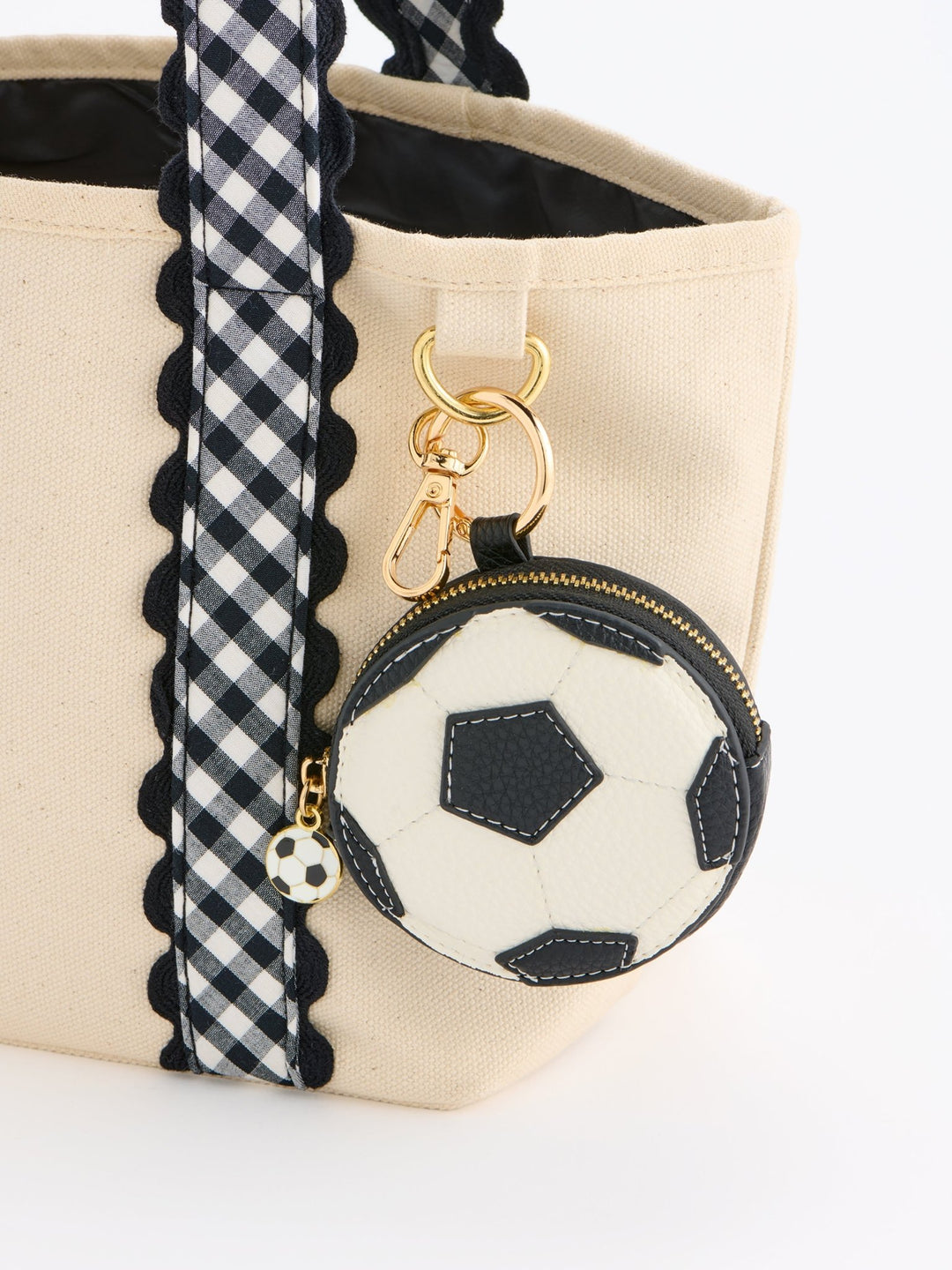 A close-up of a beige canvas tote bag with a black and white gingham patterned strap, featuring a small soccer ball-shaped zipper coin purse with gold hardware.