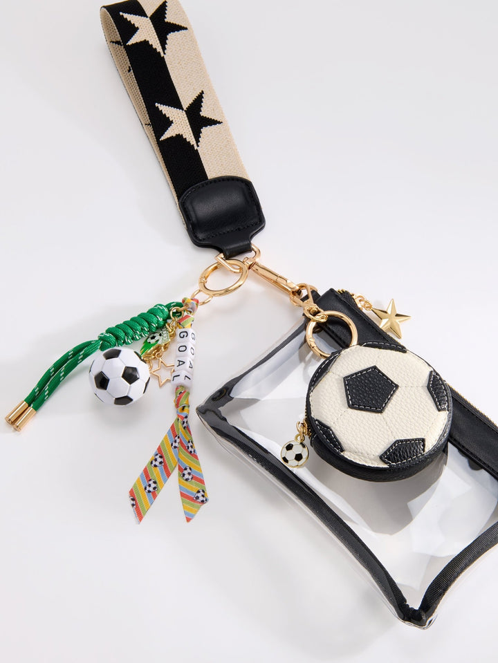 A soccer-themed clear pouch with a black strap featuring black stars, gold-tone hardware, and multiple dangling charms including a small soccer ball, a green braided tassel, and a colorful patterned ribbon.