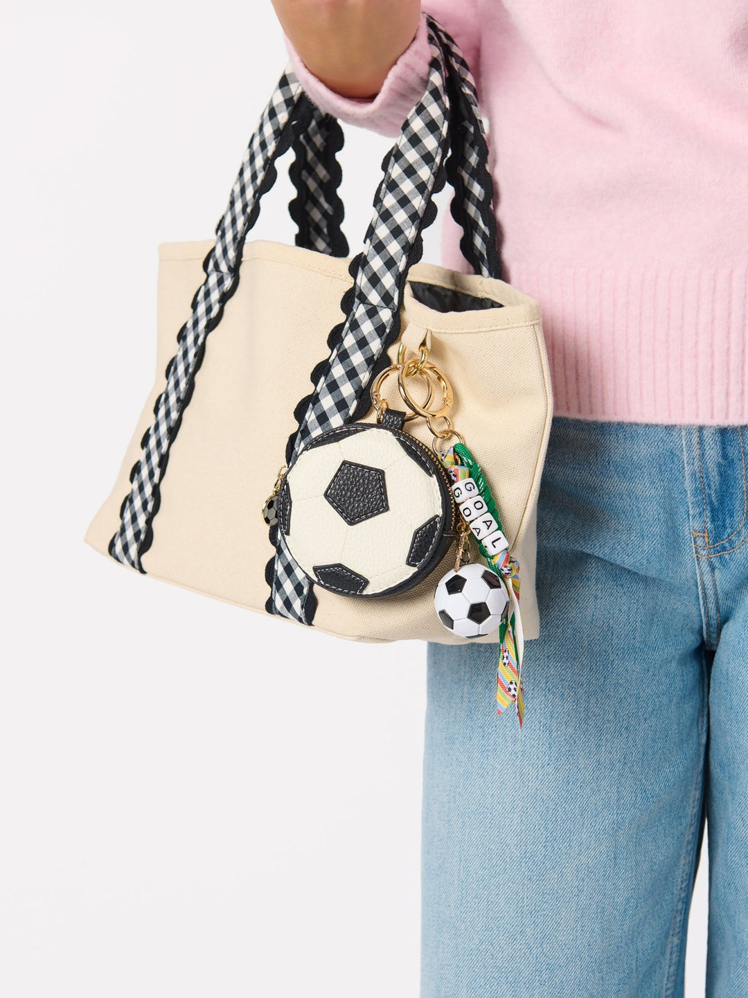 A person wearing a pink sweater and blue jeans carries a beige tote bag with black-and-white gingham braided shoulder straps and soccer-themed keychain charms.
