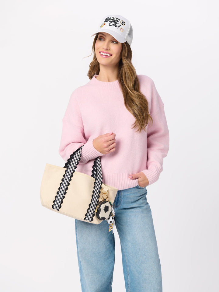 A woman modeling a light pink crewneck sweater, blue jeans, a white baseball cap, and a cream tote bag with black-and-white patterned straps, posed against a plain white studio background.