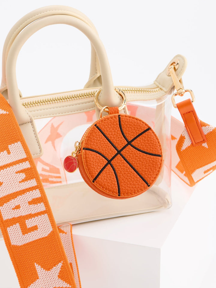 A clear acrylic handbag with cream-colored faux leather handles and trim, featuring an orange basketball-shaped coin pouch charm and an orange shoulder strap with white text, set against a clean white background.
