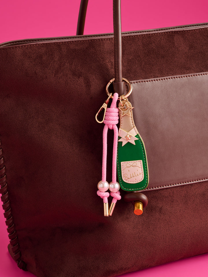 A close-up of a dark brown suede tote bag with a pink and green decorative keychain charm featuring a champagne bottle design, pearls, and gold-tone hardware, set against a bright pink background.