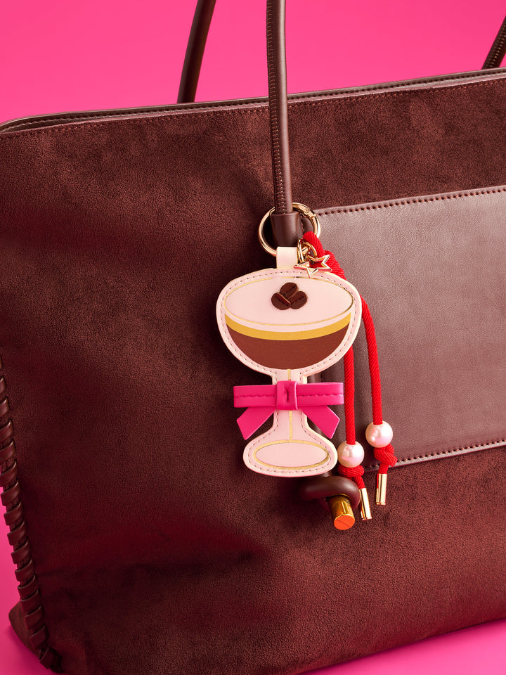 A brown suede tote bag with a matching brown leather strap, decorated with a whimsical coffee-themed keychain charm featuring a latte art design, a pink bow, and red tassels, set against a solid hot pink background.