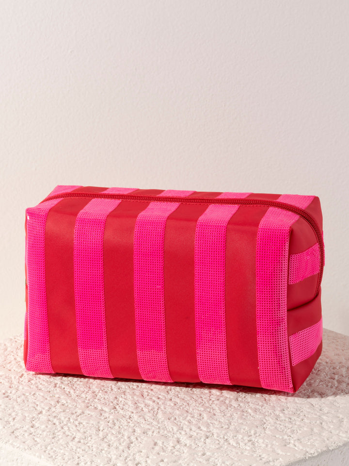 A rectangular zippered cosmetic pouch with vertical stripes in red and bright pink, resting on a textured white surface against a plain off-white wall.