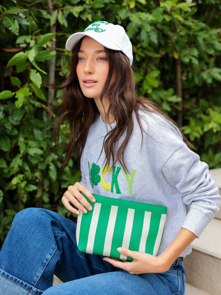 Woman wearing a gray sweatshirt with 'LUCKY' text, holding a green and white striped clutch, and wearing a white cap with green accents.