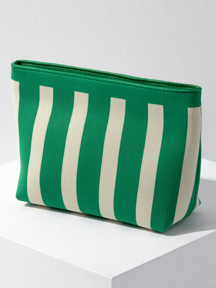 A green and off-white vertically striped canvas pouch with a zip-top closure, resting on a white geometric platform against a light gray wall.