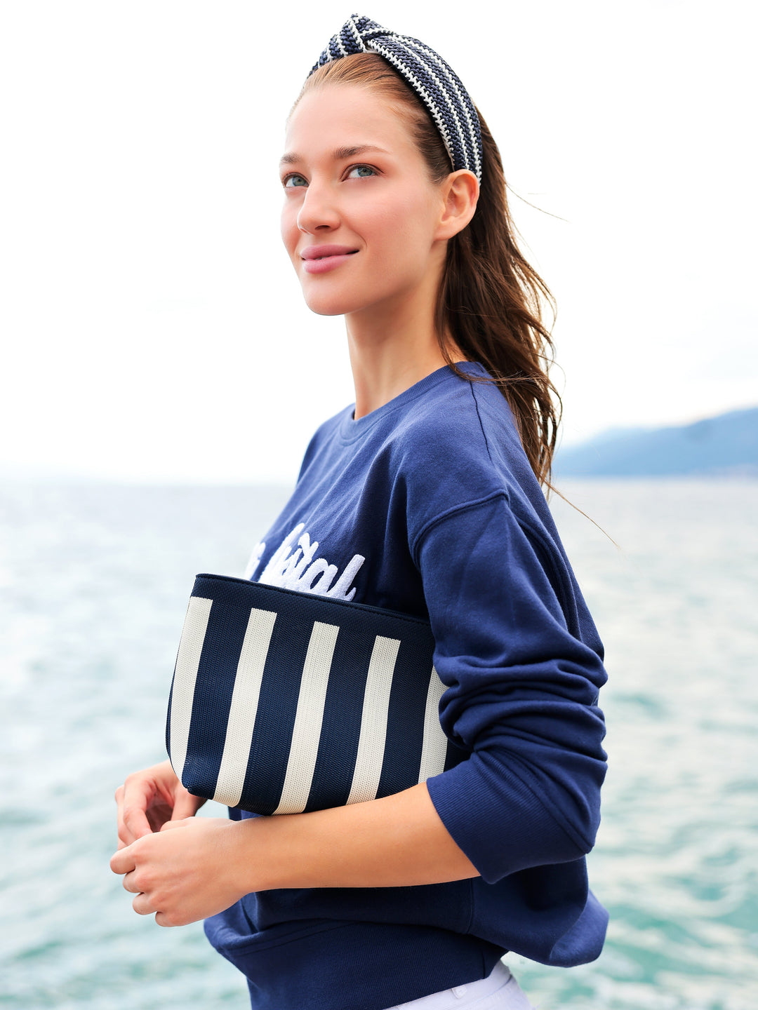 A woman wearing a navy blue sweatshirt and a black-and-white striped headband, holding a matching striped clutch bag against a blurred ocean background.