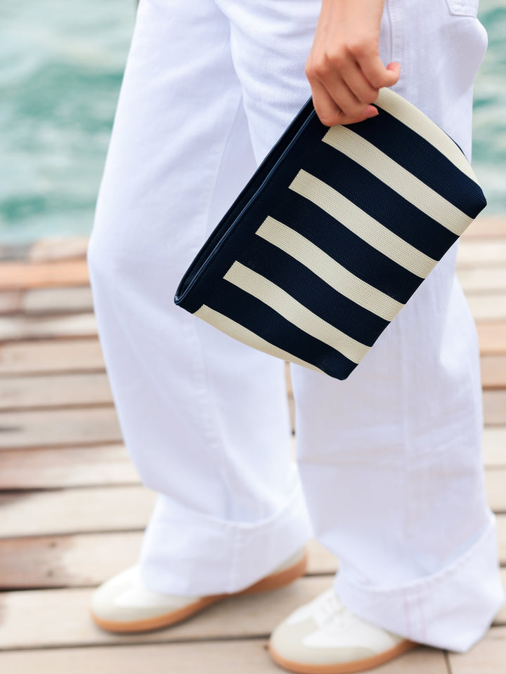 A person wearing white pants and white shoes holds a navy blue and cream striped clutch bag while standing on a wooden dock with water in the background.