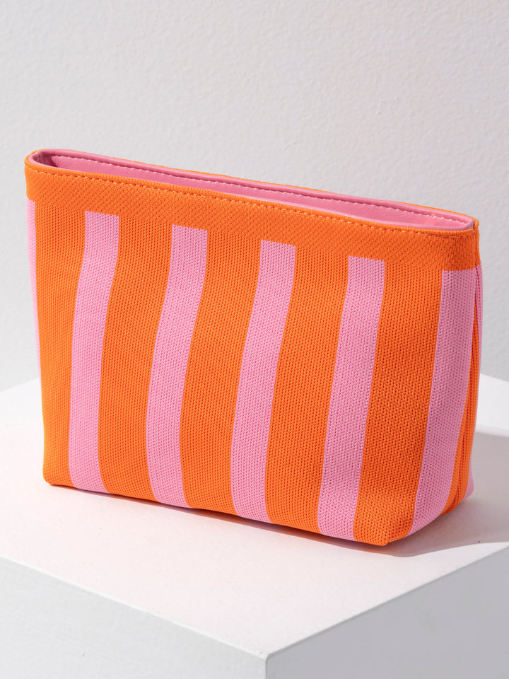An orange and pink striped zippered pouch bag resting on a white display block against a light gray wall.