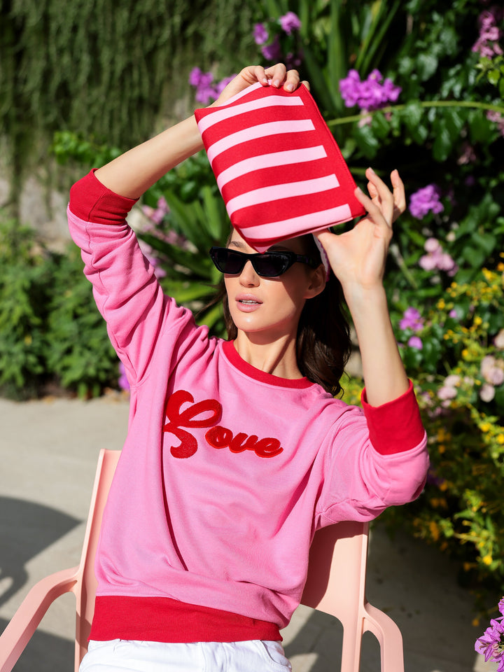 Person wearing a pink sweatshirt with 'Love' printed on it, sitting outdoors with a striped bag.