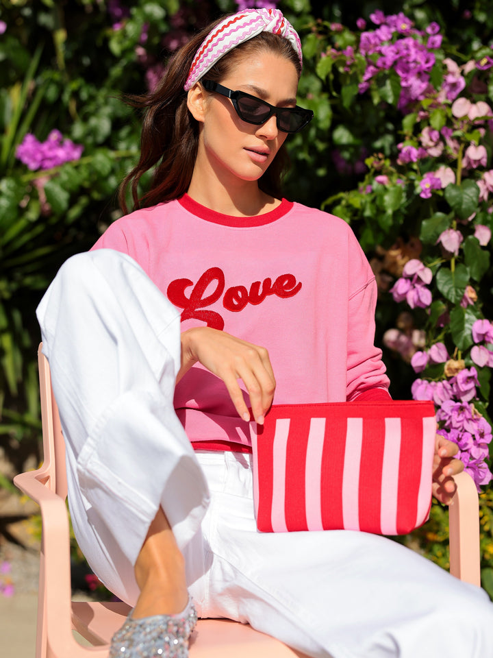 Woman in pink 'LOVE' sweatshirt and sunglasses holding a red and white striped bag, sitting outdoors with flowers in the background.