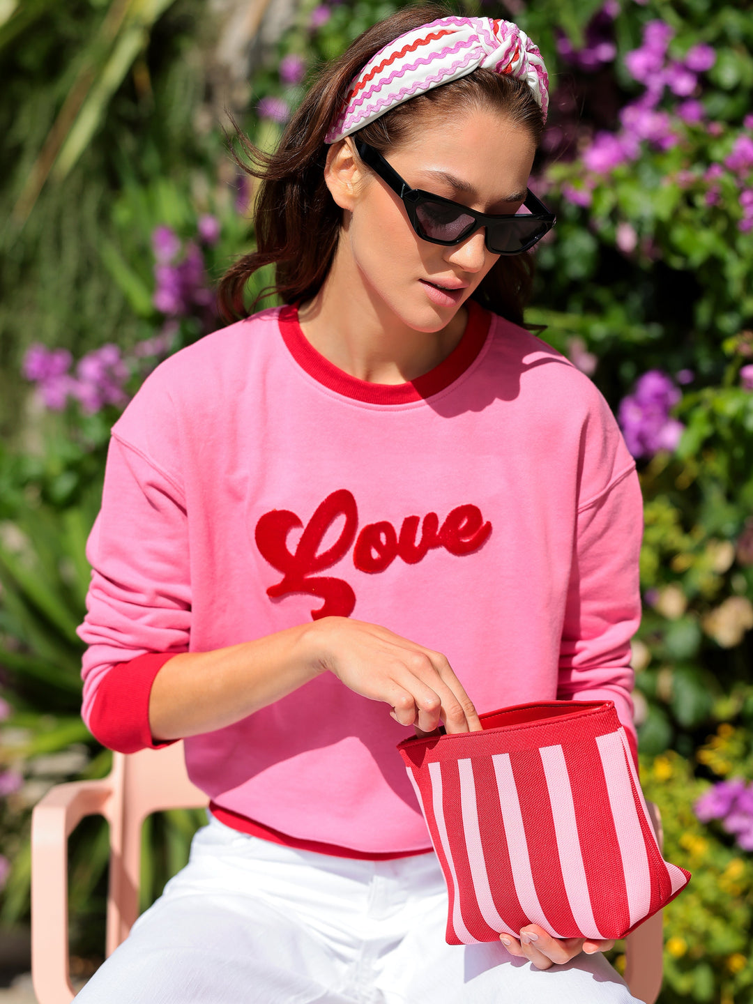 Woman wearing a pink sweatshirt with 'Love' on it, holding a red and white striped bag, sitting outdoors.
