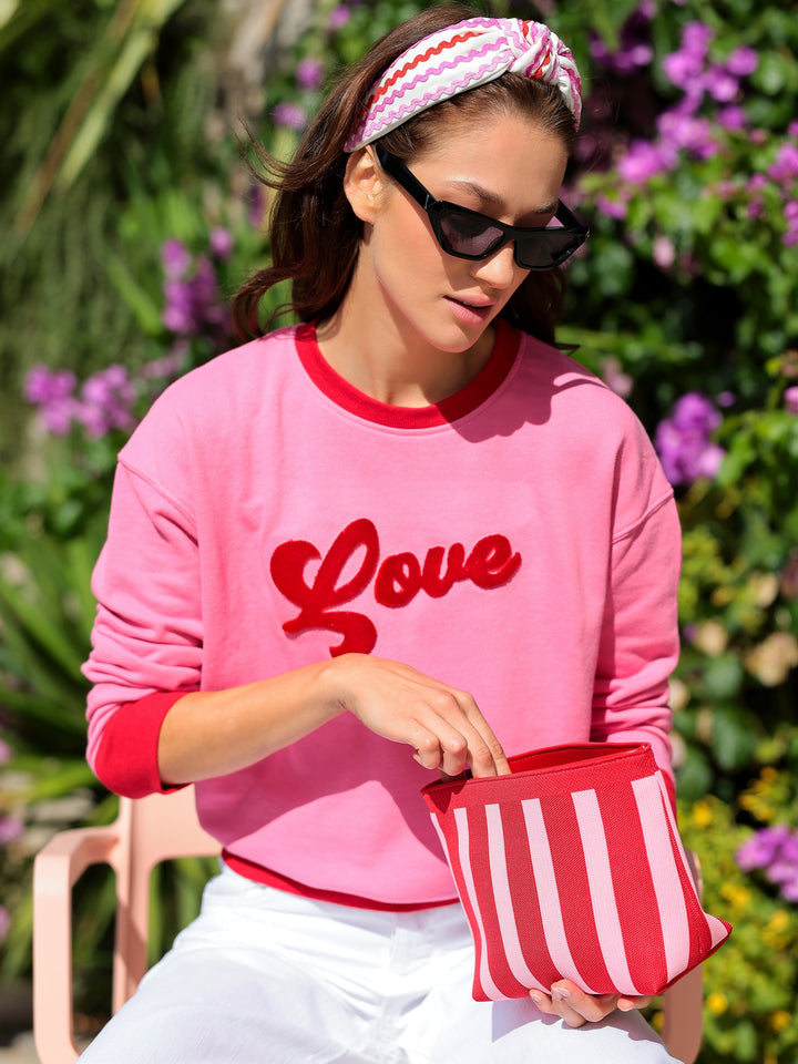 Woman wearing a pink sweatshirt with 'Love' on it, holding a red and white striped bag, sitting outdoors.