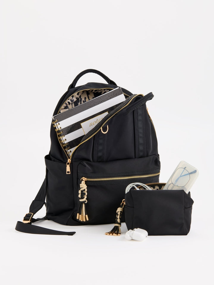 A black nylon backpack with gold-tone hardware and tassel zipper pulls, shown open with a patterned interior and packed with notebooks, alongside a matching black pouch and small accessories on a clean white background.