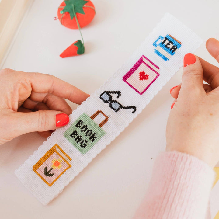 Hands holding the bookish cross stitch bookmark beside a tomato pincushion