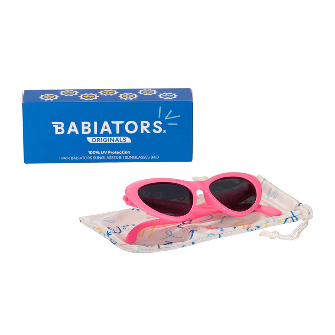 Pink cat-eye kids sunglasses folded on a printed storage pouch beside the Babiators Originals box.