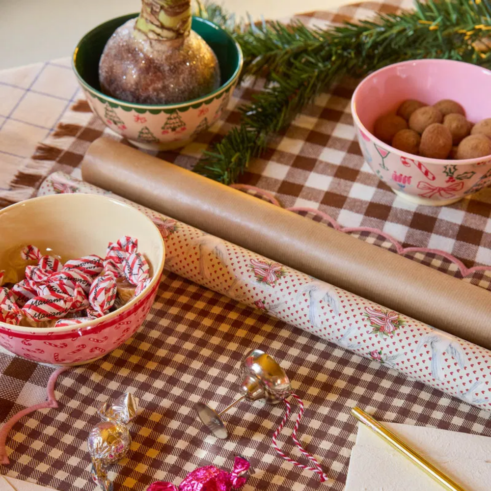 Candy cane bowl full of peppermints beside other Christmas bowls on brown gingham with a roll of wrapping paper