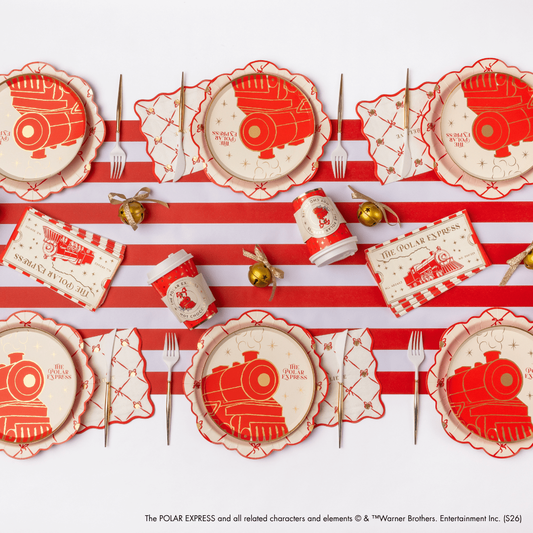 A festive holiday tableware set featuring red and cream paper plates, napkins, cups, and placemats with a vintage train illustration, arranged on a red and white striped background with gold cutlery and small gold bells.