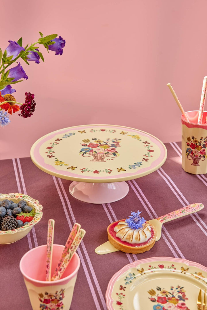 Nathalie Lété cake stand on a striped tablecloth with berries, floral cups, and plates against a pink wall
