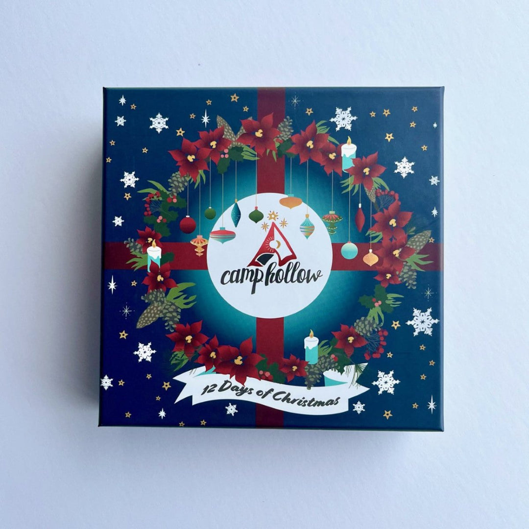 A square Christmas advent box by CampHollow, featuring a dark blue background decorated with a red and green wreath, red poinsettias, white snowflakes, and a white circular logo in the center.