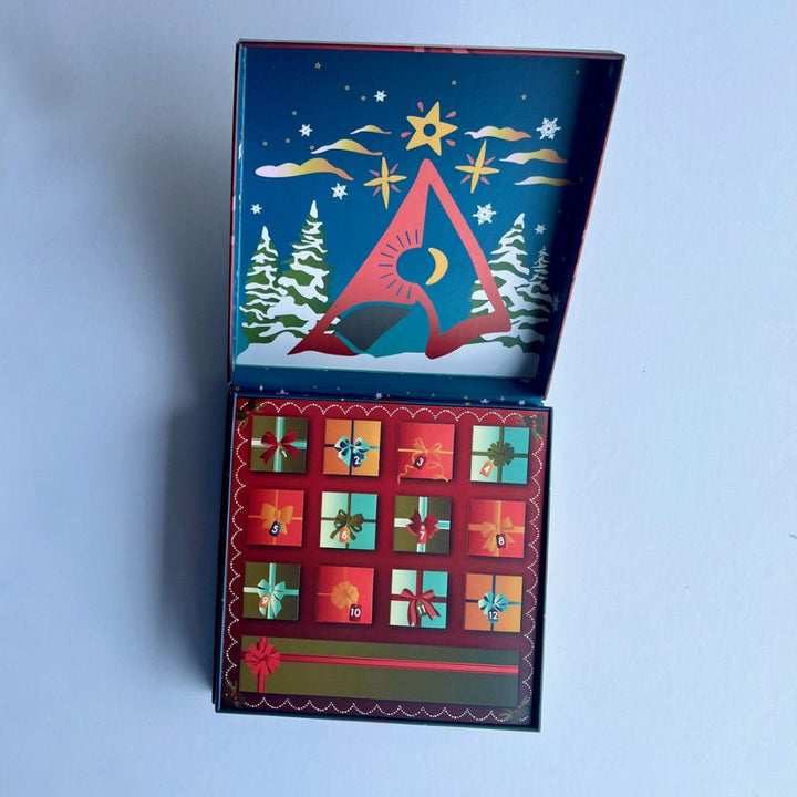 An open Christmas advent calendar box with a winter night scene illustration on the inside lid and 12 advent pockets decorated with colorful gift boxes on the front, set against a light blue background.