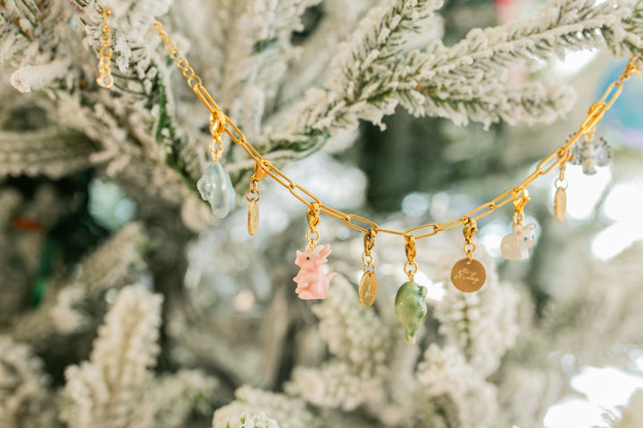 A gold holiday garland with small pastel charms, including a pink bunny, green teardrop, and clear beads, hanging against a blurred flocked Christmas tree background.